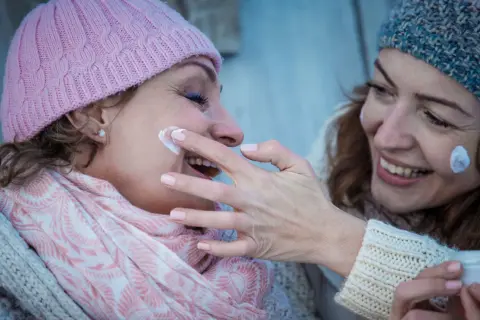 Dos mujeres sonríen mientras se aplican crema. Una tiene la piel alipica y lleva un gorro rosa, la otra, un gorro azul.