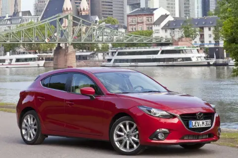 Czerwony sedan Mazda 3 na tle miasta z mostem i rzeką.