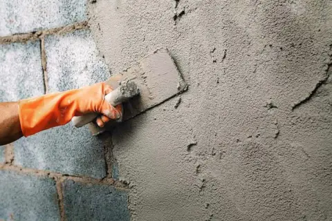 Mano con guante naranja aplica enfoscado rugoso a pared de bloques de cemento con llana.