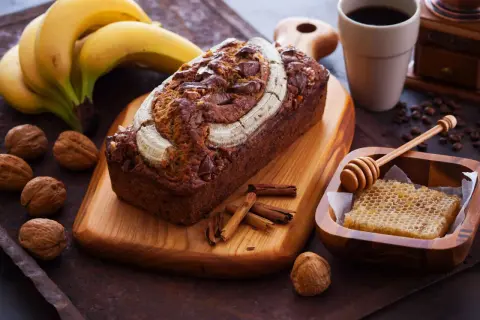 Ein saftiges Bananenbrot mit Schokoladenstückchen und Nüssen, garniert mit einer Banane. Daneben Kaffee, Honigwabe und Zimtstangen – ein Genuss, der auch bananenbrot gesund macht.