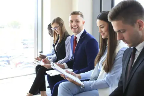 Giovani professionisti in attesa per un colloquio di gruppo, alcuni con appunti, uno sorride.