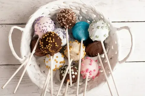 Kolorowe cake pops w białym koszyku, udekorowane posypkami i kuleczkami.