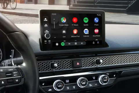 Wnętrze samochodu z ekranem Android Auto, na którym widać ikony Google Maps, Spotify i innych aplikacji. W tle dyskretnie widać Yanosik.
