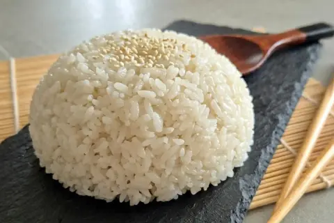Ein Berg aus weißem Reis, bestreut mit Sesam, auf einem schwarzen Stein. Daneben liegen Stäbchen und ein Holzlöffel. Das ist die Basis, was bedeutet Sushi.