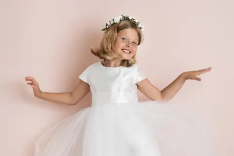 A smiling young girl in a white dress and flower crown, with arms outstretched.