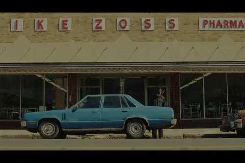 Niebieski samochód przed apteką "MIKE ZOSS PHARMACY". Obraz w stylu **false color**, z lekko zmienionymi barwami, tworzy nostalgiczny klimat.
