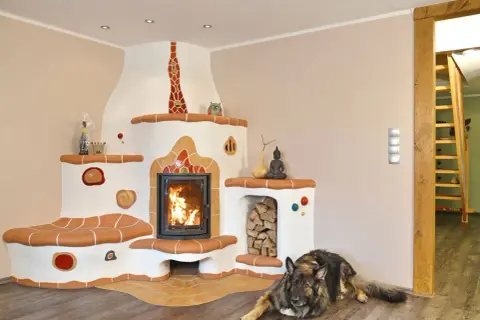 Ein Hund liegt vor einem großen, weißen Kachelofen mit integriertem Kamin. Der Ofen hat eine Sitzbank und ist mit bunten Kacheln verziert. Der Unterschied zwischen Kamin und Ofen wird hier durch die massive Bauweise und die Heizfunktion des Ofens deutl...