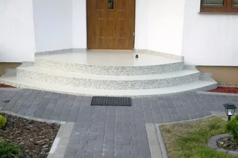 Nowe schody z żywicy na balkon, z kamyczkami, prowadzące do drewnianych drzwi. Wokół kostka brukowa i roślinność.