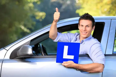 Hombre sonriente en coche con cartel "L", pulgar arriba. Pregunta: ¿cuanto tiempo hay que llevar la L?