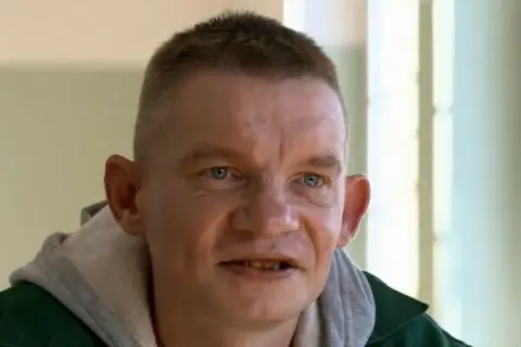 Mężczyzna z krótkimi włosami i niebieskimi oczami, ubrany w zieloną kurtkę i szary kaptur. Wygląda jakby miał zarost, a jego usta są lekko otwarte. Może to nasz bandziorek?
