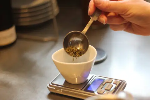 Une main verse des herbes séchées dans une tasse blanche posée sur une balance. Le dosage précis est essentiel pour 1 litre d'infusion.