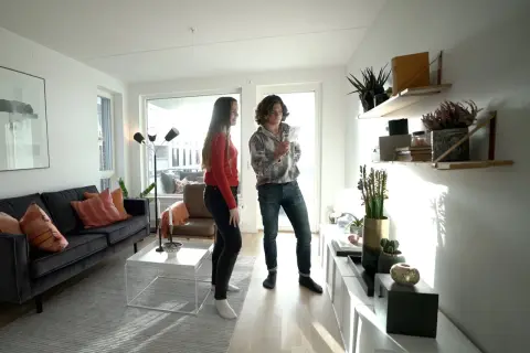 Två personer i ett ljust vardagsrum med stilren inredning. En mörk soffa med kuddar, ett vitt soffbord och en golvlampa. Hyllor med växter och dekorationer.