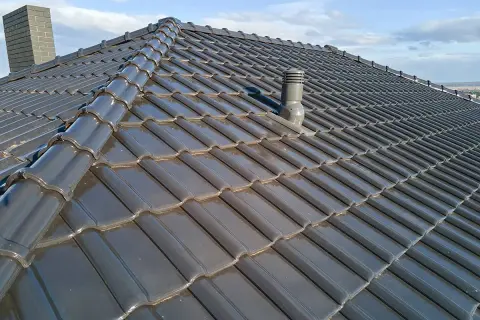 Szary dach z dachówki ceramicznej, z widocznym kominkiem wentylacyjnym i kominem. Rozmieszczenie kominków wentylacyjnych na dachu zapewnia prawidłową cyrkulację powietrza.