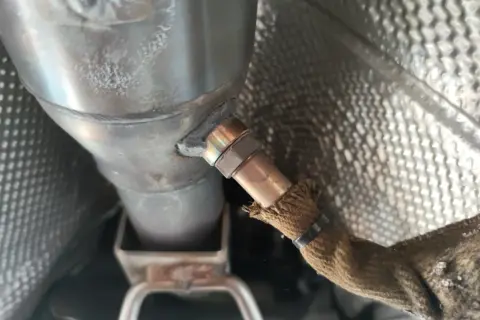 Nahaufnahme einer Lambdasonde, die am Auspuffrohr eines Autos befestigt ist. Sie misst den Sauerstoffgehalt im Abgas, um die optimale Verbrennung zu gewährleisten.