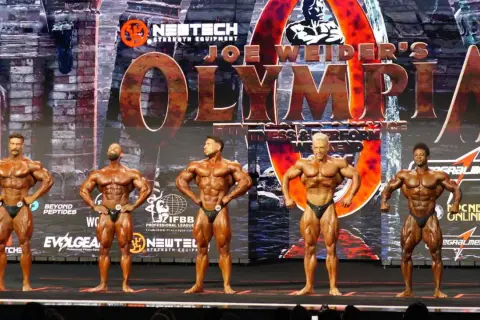 Fünf muskulöse Männer posieren auf der Bühne bei einem Bodybuilding-Wettkampf. Die "Joe Weider's Olympia" Bühne ist im Hintergrund zu sehen.