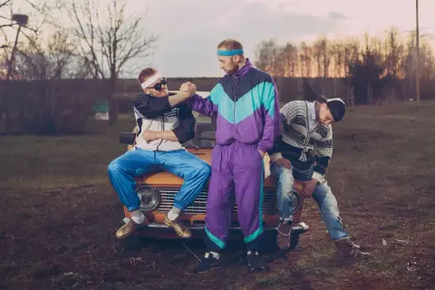 Trzech mężczyzn w dresach w stylu lat 90. siedzi na masce starego samochodu.