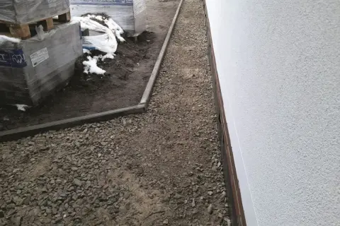 Ułożono kruszywo kamienne tłuczeń wzdłuż białego muru, obok palet z materiałami budowlanymi.