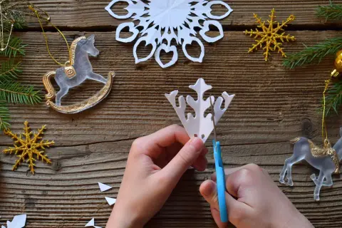 Ręce tnące papierową śnieżynkę na drewnianym tle z ozdobami świątecznymi.