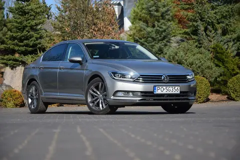 Szary Volkswagen Passat sedan zaparkowany na podjeździe, otoczony zielenią i kamieniami.
