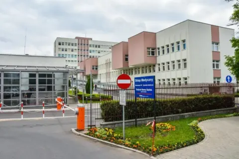Nowoczesny budynek szpitala z bramkami wjazdowymi i znakami informacyjnymi.