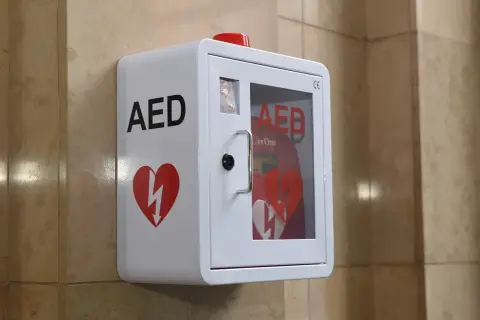 Biała szafka AED z czerwonym sercem i błyskawicą, zamontowana na ścianie.