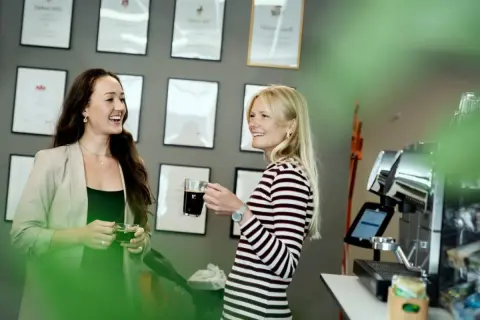Två kvinnor, en grafisk formgivare, pratar vid en kaffemaskin. Väggen bakom dem är prydd med diplom.