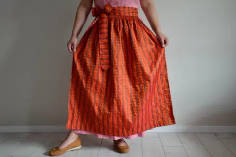 Eine Frau trägt ein Dirndl mit einer leuchtend orangefarbenen Dirndlschürze, die mit Streifen und kleinen Tupfen gemustert ist.
