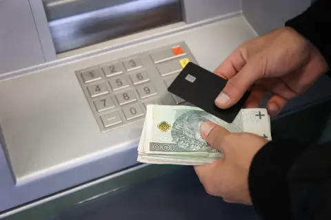 Ręce trzymające banknoty i kartę zbliżają się do wpłatomatu. Pieniądze na koncie czekają na wpłatę.