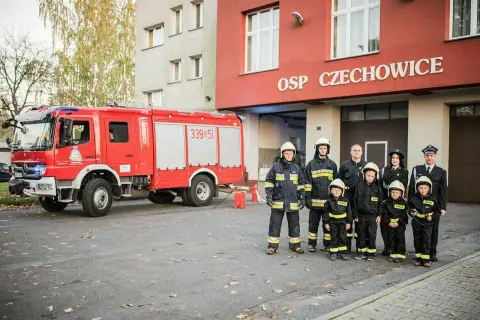 Strażacy OSP Czechowice z wozem strażackim i dziećmi w strojach strażackich.