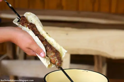 Ręka trzyma lula kebab zawinięty w placek. Mięso jest soczyste i rumiane, gotowe do jedzenia.