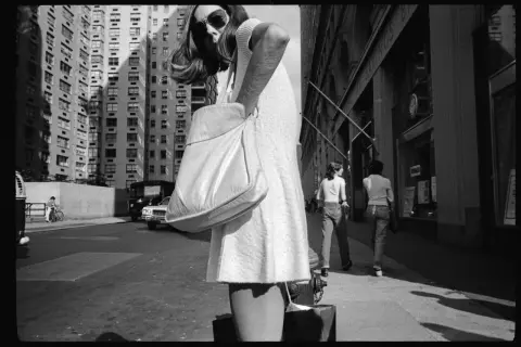 Una donna con borsa a tracolla cammina per strada, con palazzi alti sullo sfondo. Mark Cohen photography cattura la vita urbana.