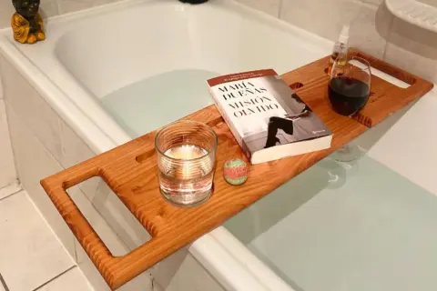Tabla de madera sobre bañera con libro, vaso de agua, bomba de baño y copa de vino. Preparando un baño relajante.