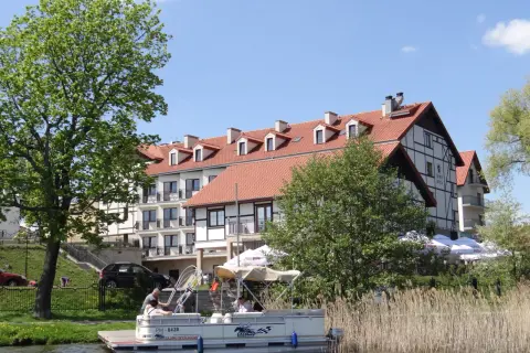 Hotel Anek Mrągowo nad jeziorem, z łodzią przy pomoście i zielenią.