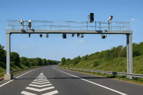 Portique de péage sans barrière sur une autoroute bordée d'arbres. Des caméras et des feux sont installés sur la structure métallique.
