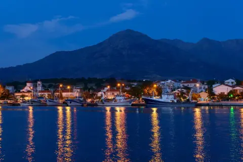 Port z samotraki nocą. Łodzie cumują przy nabrzeżu, a światła odbijają się w spokojnej wodzie. W tle widać góry.