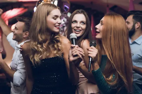 Trzy kobiety śpiewają do mikrofonów, bawiąc się przy karaoke. W tle widać innych ludzi tańczących.