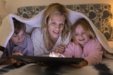 Mama z dziećmi pod kocem ogląda bajkę na tablecie. To piękny przykład, jak wychować dziecko w bliskości i wspólnocie.