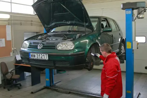 Mechanik w czerwonej kurtce sprawdza podniesiony na podnośniku zielony samochód. To kluczowy moment, by dowiedzieć się, jak dbać o samochód.