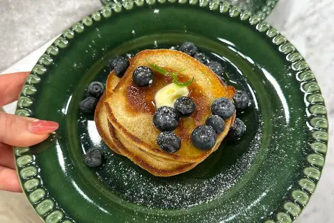 En hög med amerikanska pannkakor toppas med smör, blåbär och sirap på en grön tallrik.