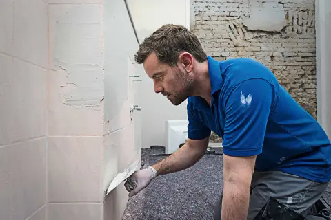 Mann spachtelt Fliesen mit einer Kelle. Er trägt Handschuhe und ein blaues Polohemd.
