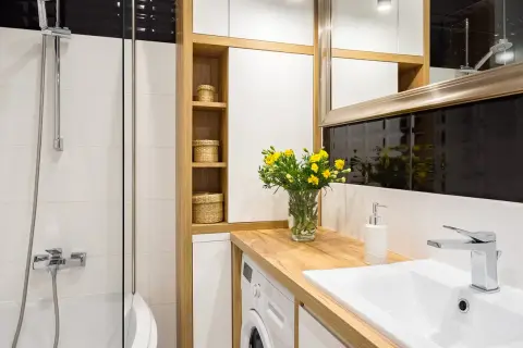 Petite salle de bain avec baignoire, lavabo, machine à laver et étagères en bois. Un bouquet de fleurs jaunes décore le plan de travail.
