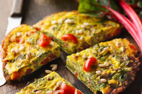 Tortilla de acelgas con tomates cherry y piñones, cortada en porciones sobre una tabla de madera.