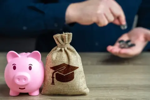 Różowa świnka-skarbonka i worek z symbolem czapki absolwenta na drewnianym stole.