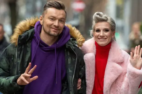 Sylwia Grzeszczak i Liber uśmiechają się do obiektywu, ona macha ręką, on pokazuje znak pokoju.