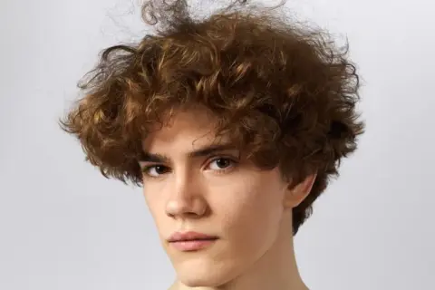 Primer plano de un joven con cabello rizado y castaño sobre fondo blanco.