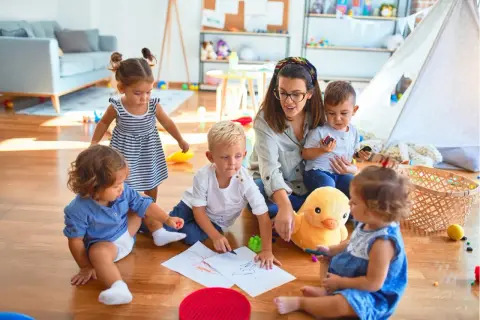 Eine Tagesmutter wird, umgeben von spielenden Kindern. Sie malt mit ihnen auf dem Boden, während ein Stoffentier zusieht.