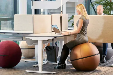 Kvinna arbetar vid ett höj- och sänkbart skrivbord i ett aktivitetsbaserat kontor. Hon sitter på en stor, brun träningsboll.