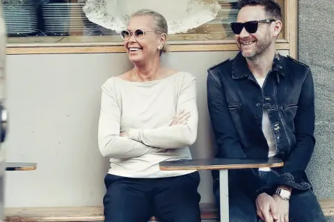 Joakim Berg familj: en kvinna och en man sitter på en bänk utanför ett café, båda ler och bär solglasögon.