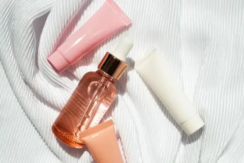 Un frasco de serum con gotero y tubos de crema rosa, blanco y melocotón sobre tela blanca. ¿Que va primero el serum o la crema?