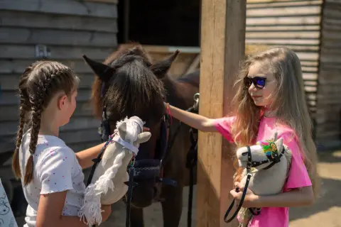 Dwie dziewczynki bawią się pluszowymi końmi. Jedna z nich głaszcze prawdziwego konia, a druga trzyma swojego hobby horse.